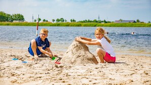 Dos niños construyen un castillo de arena en la playa, con agua tranquila y vegetación al fondo.