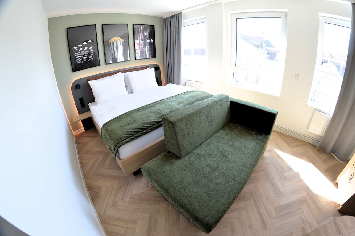 Une chambre d’hôtel moderne, dotée d’un lit, d’un banc recouvert d’un tissu vert et de grandes fenêtres offrant une vue sur la ville.