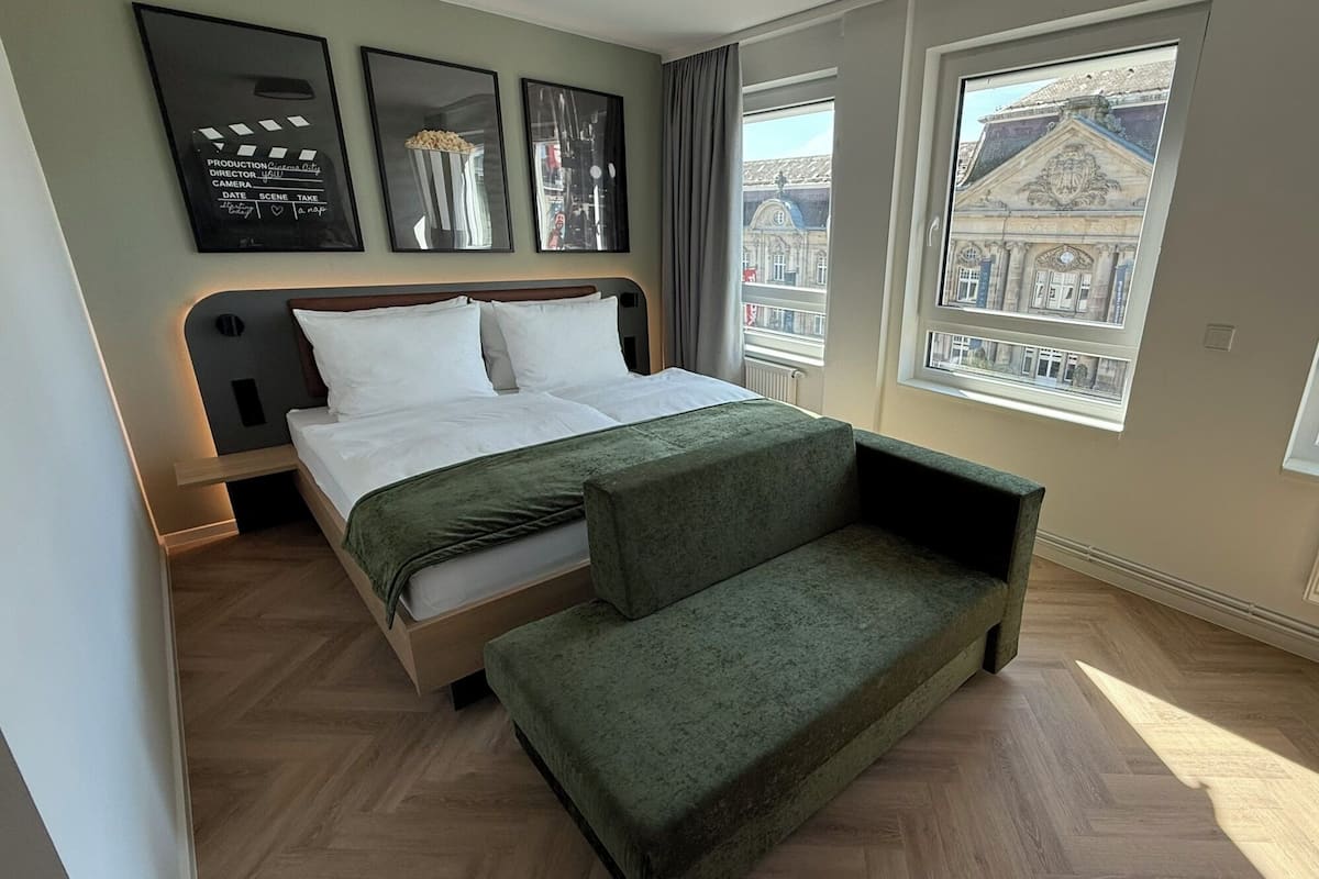Une chambre moderne avec un lit, un canapé vert et une grande fenêtre donnant sur un bâtiment historique.