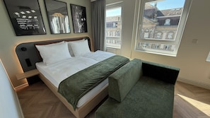 A modern hotel room with a neatly made bed, a green upholstered bench, and a large window offering a view of historic buildings. - Cinema City Hotel Europaplatz Karlsruhe (Karlsruhe)