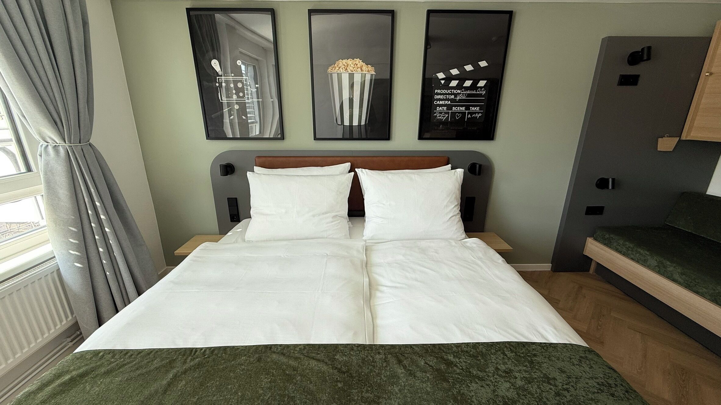 Un lit bien fait, avec du linge de lit blanc, une tête de lit ornée d’un panneau marron et un plaid vert au pied du lit. Trois tableaux encadrés sont accrochés au mur, au-dessus du lit.