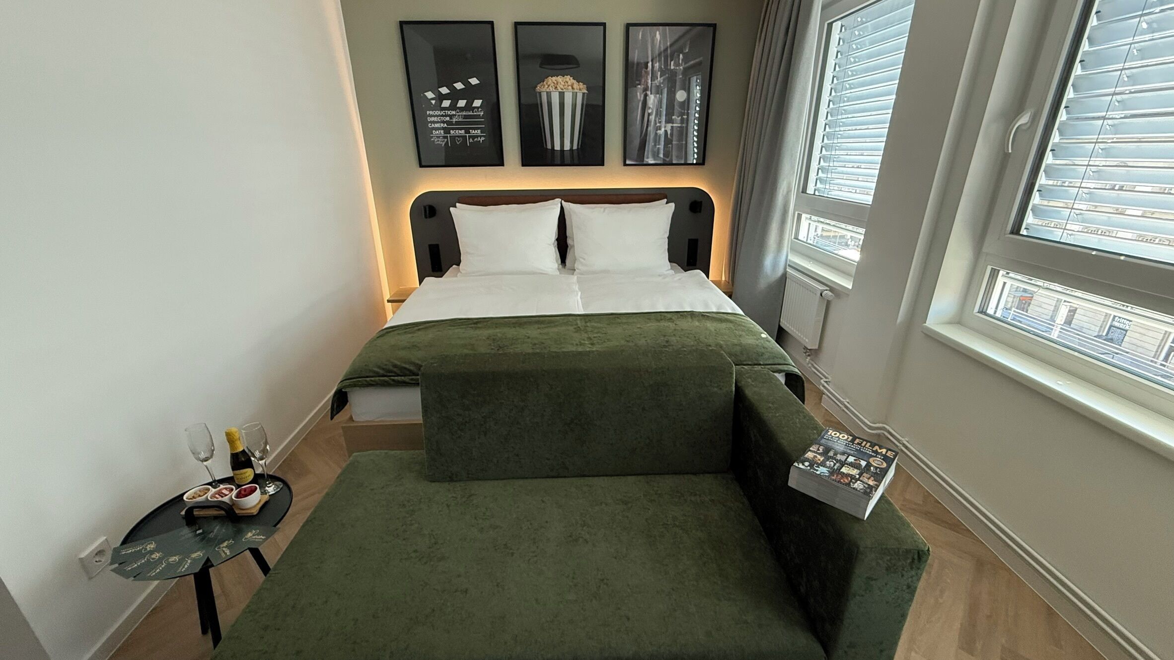 A hotel room with a bed, a small table with a bottle and glasses, a magazine, and a window with blinds.