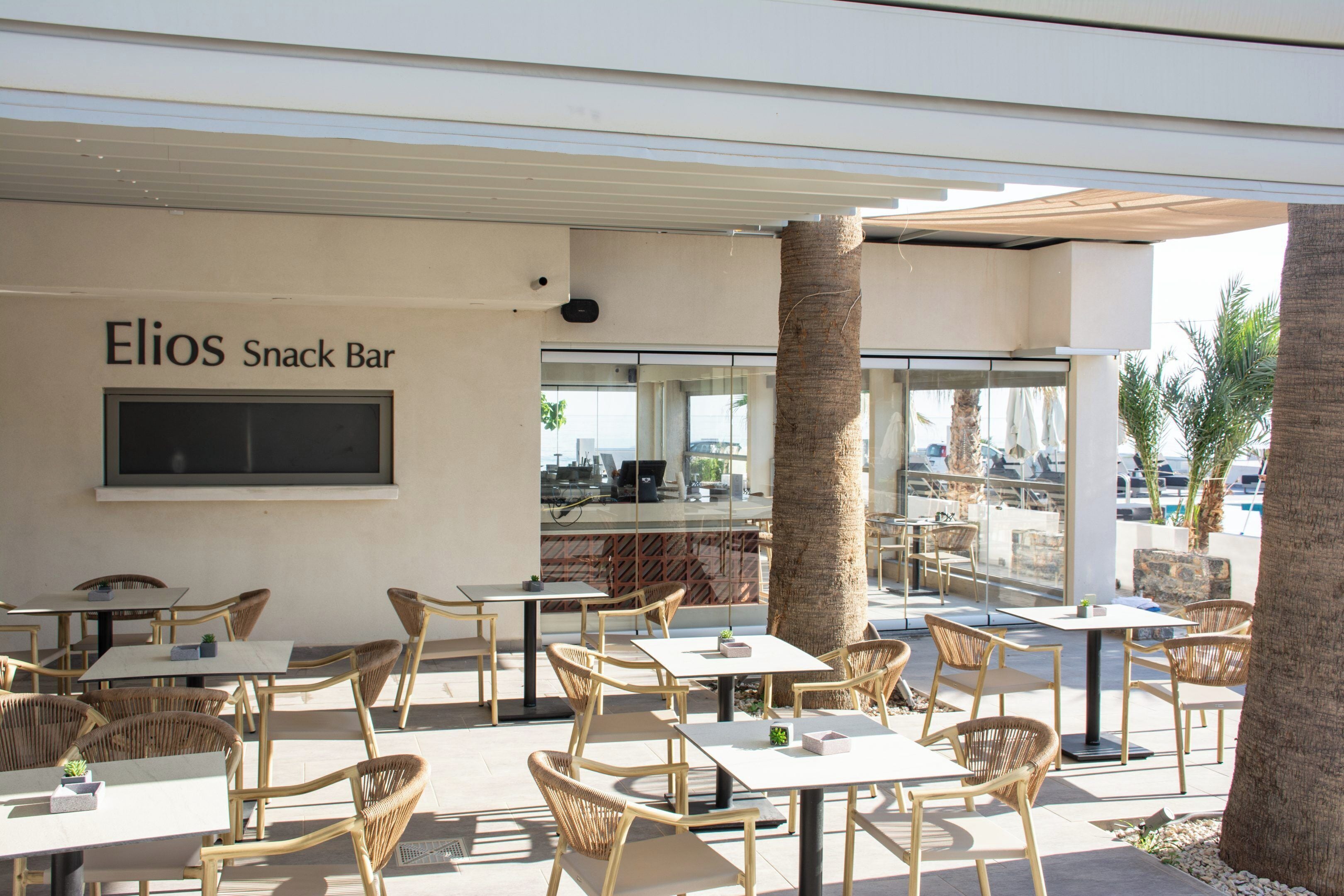 Ein Sitzbereich im Freien mit Korbstühlen und Tischen, ein Schild für 'Elios Snack Bar' und im Hintergrund eine Ansicht der Straße und der Gebäude.