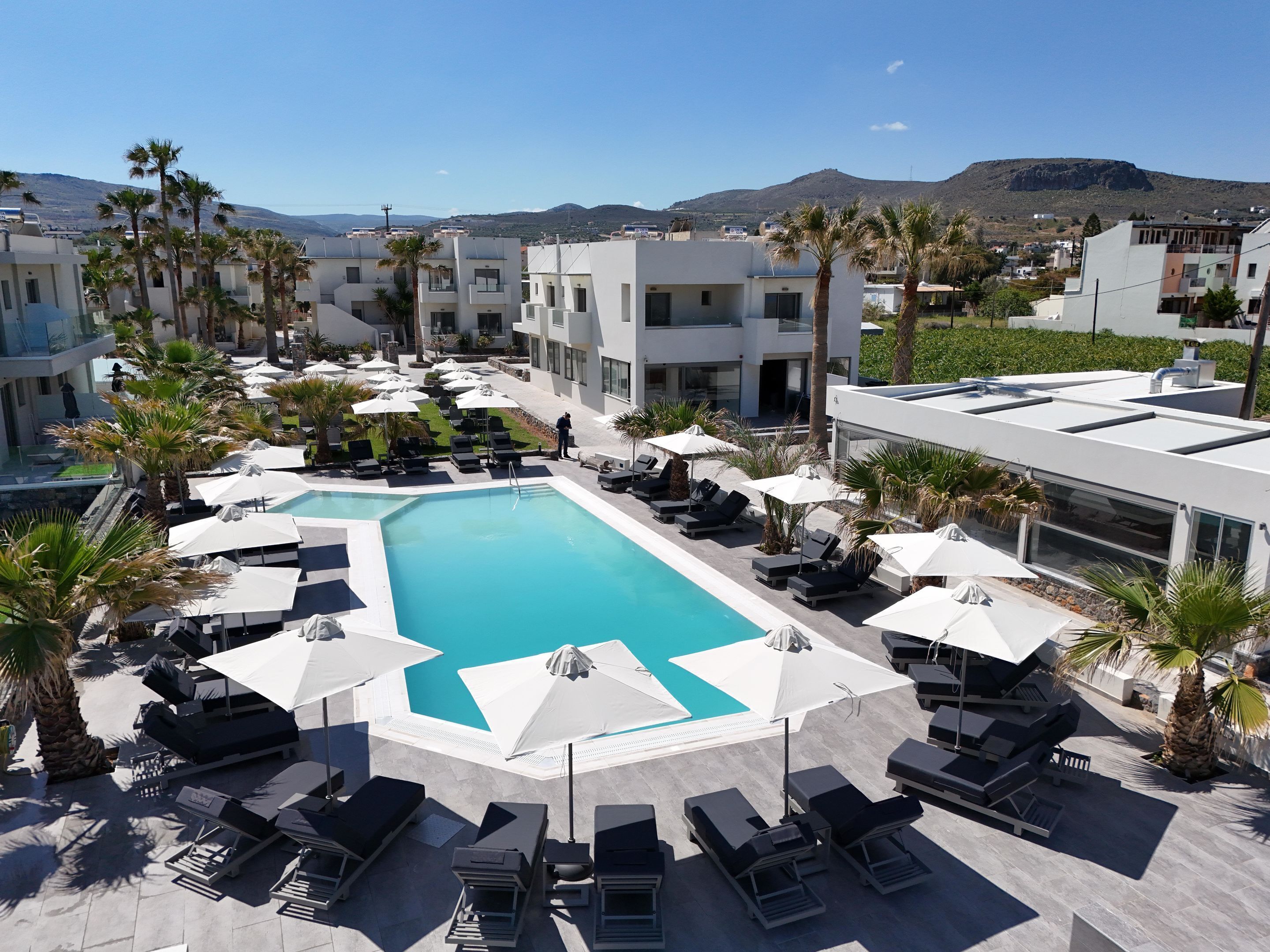 A resort pool area with sun loungers and umbrellas, surrounded by palm trees and modern buildings.