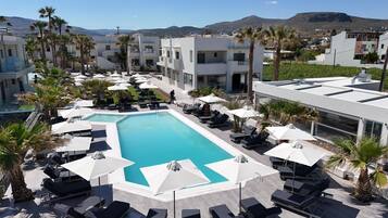 A resort pool area with sun loungers and umbrellas, surrounded by palm trees and modern buildings.