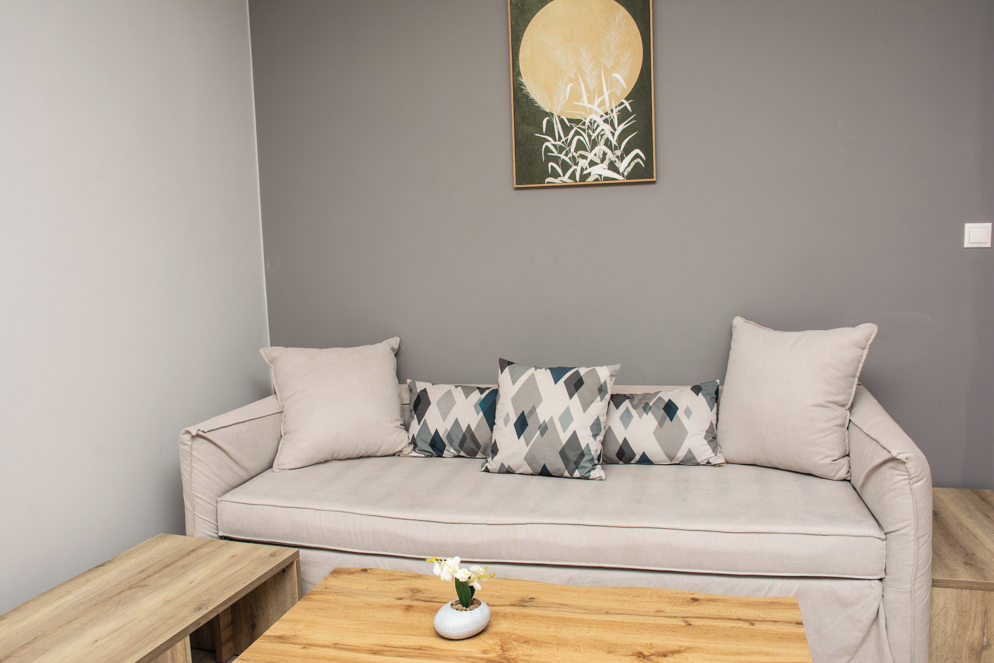 A neutral-toned sofa with patterned pillows, a wooden coffee table with a vase of flowers, and a wall-mounted artwork.