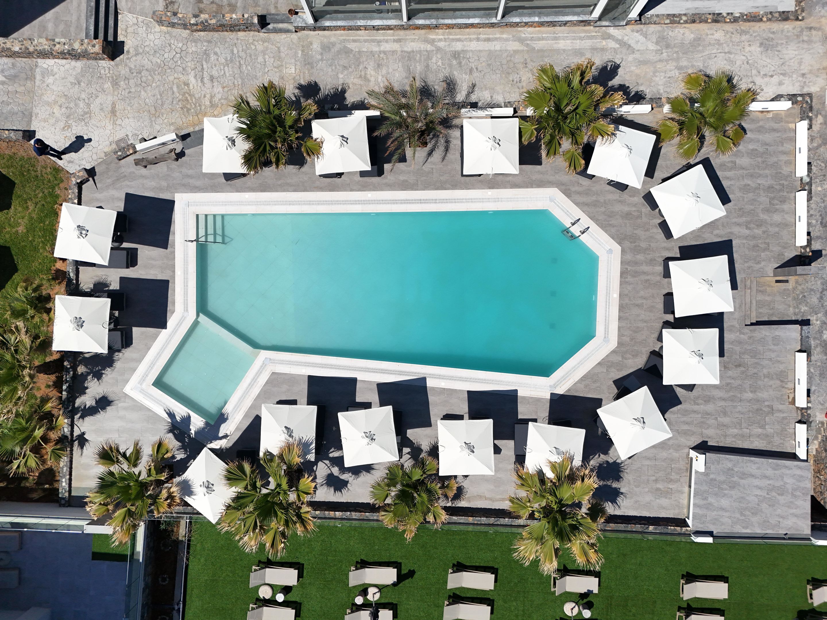 An aerial view of a swimming pool surrounded by white umbrellas and lounge chairs, with palm trees and a stone pathway.