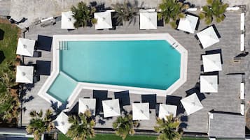An aerial view of a swimming pool surrounded by white umbrellas and lounge chairs, with palm trees and a stone pathway.