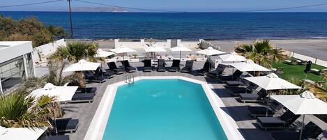 Un espace piscine doté de chaises longues et de parasols, avec vue sur une plage et l’océan.