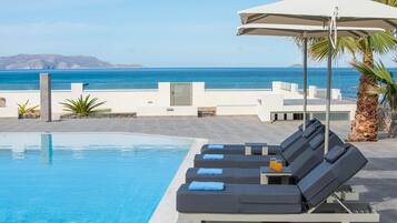 Ein Poolbereich mit Liegestühlen und einem Tisch, eine Palme und Blick auf das Meer.