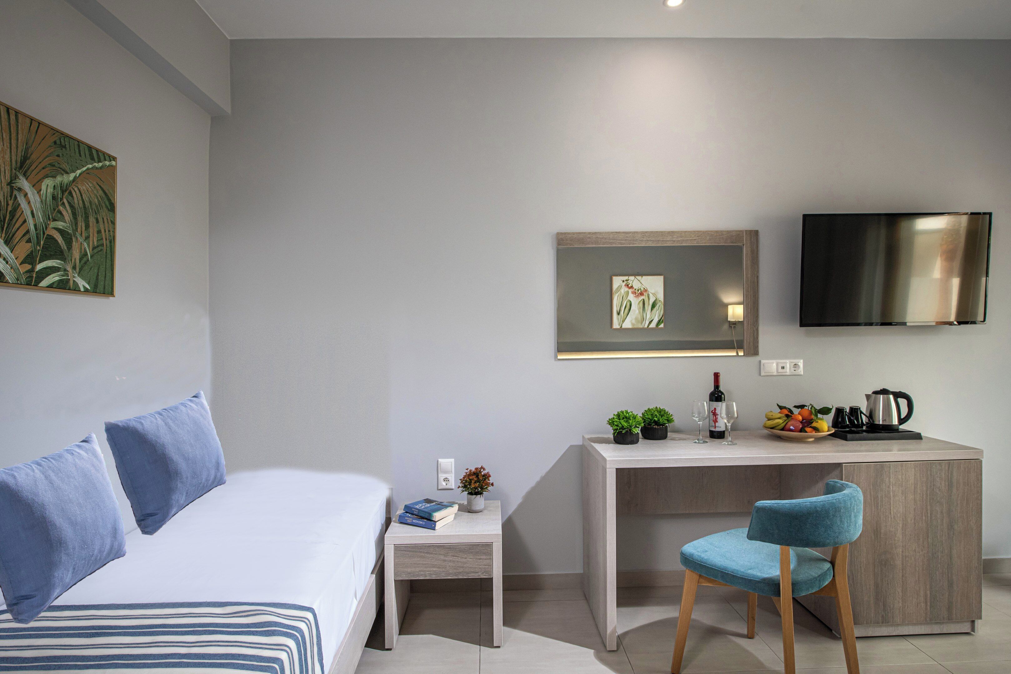 A modern hotel room with a bed, a desk, a chair, a TV, and a small kitchen area.