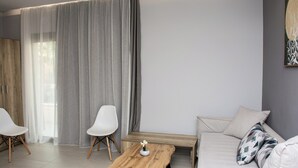 Un salon moderne avec un canapé, deux fauteuils blancs, une table basse en bois et une grande fenêtre agrémentée de rideaux gris.