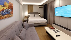 A modern hotel room with a bed, a gray sofa, a flat-screen TV, and a painting on the wall.