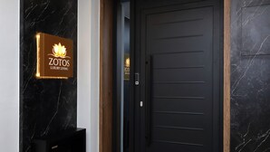 A black door with a keypad lock, a sign with a lotus flower and the text "ZOTOS LUXURY LIVING," and a dark marble wall with white veins.