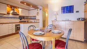 A modern kitchen with white cabinets, a wooden dining table set for four, and a red upholstered chair. - Beach and Comfort Your Stay Near the Sea (Valras-Plage)