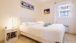 A neatly made bed with a white headboard, a bedside lamp, a small bedside table with books, and a window with bars. - Beach and Comfort Your Stay Near the Sea (Valras-Plage)