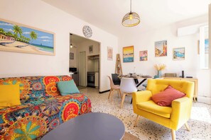 Una sala de estar con un sofá colorido, un sillón amarillo, una mesa de comedor con sillas y una cocina al fondo.