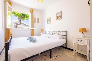 A bedroom with a bed, a window with curtains, a bedside table with a lamp, and a wall with framed pictures. - Title Valras-Loc Cosy Apartment 150m from the Beach (Valras-Plage)