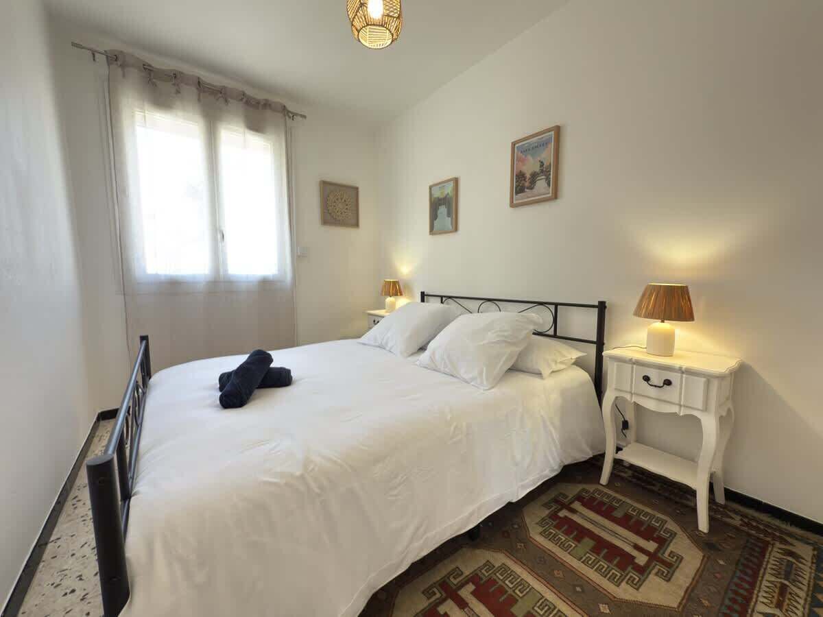 Cama bem arrumada com lençóis brancos, mesa de cabeceira com abajur e tapete estampado no chão.