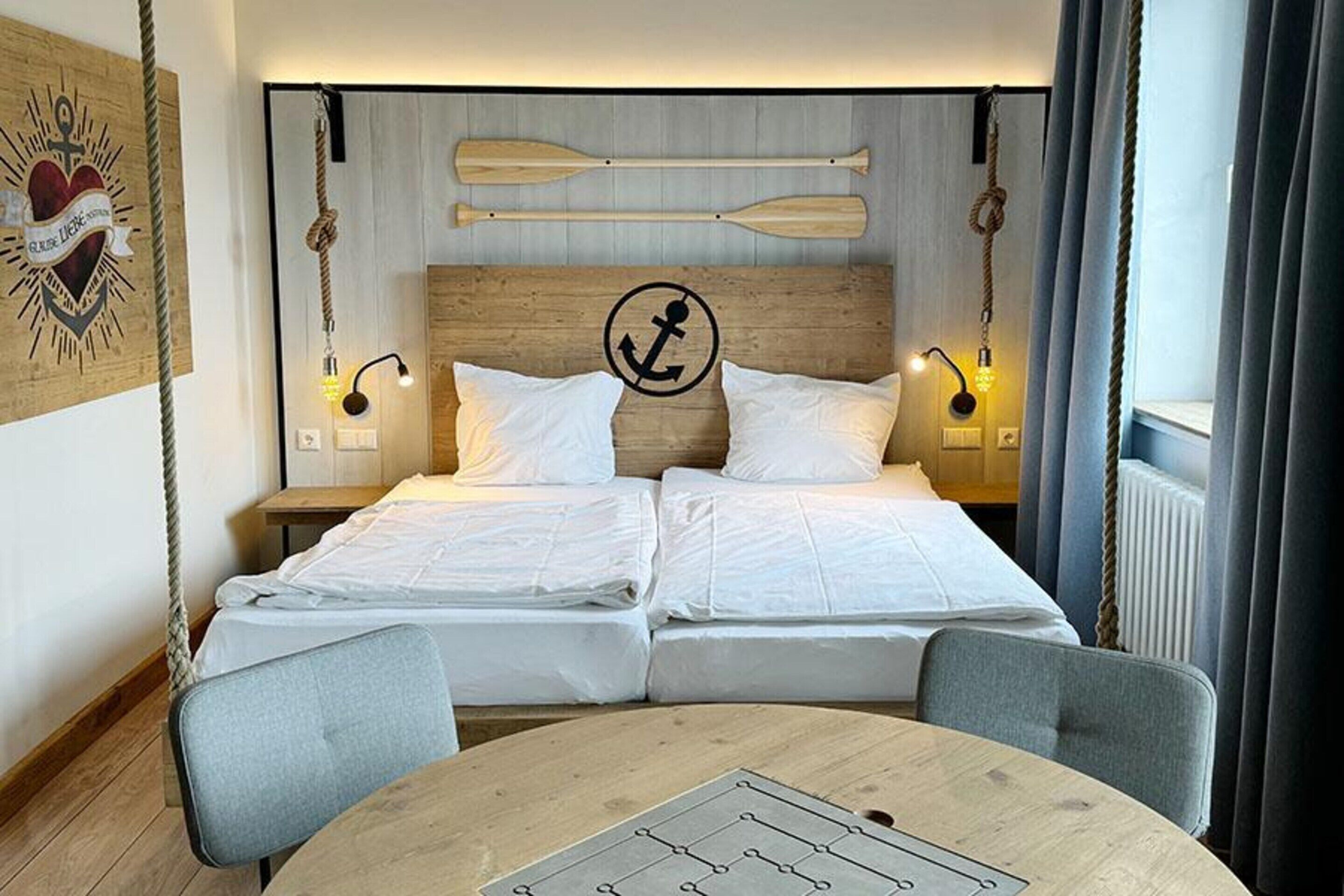 Ein Doppelbett mit weißer Bettwäsche, ein hölzernes Kopfende mit Anker-Symbol, zwei Nachttische mit Lampen, ein runder Tisch mit Schachbrett und eine Seil-Wanddekoration.
