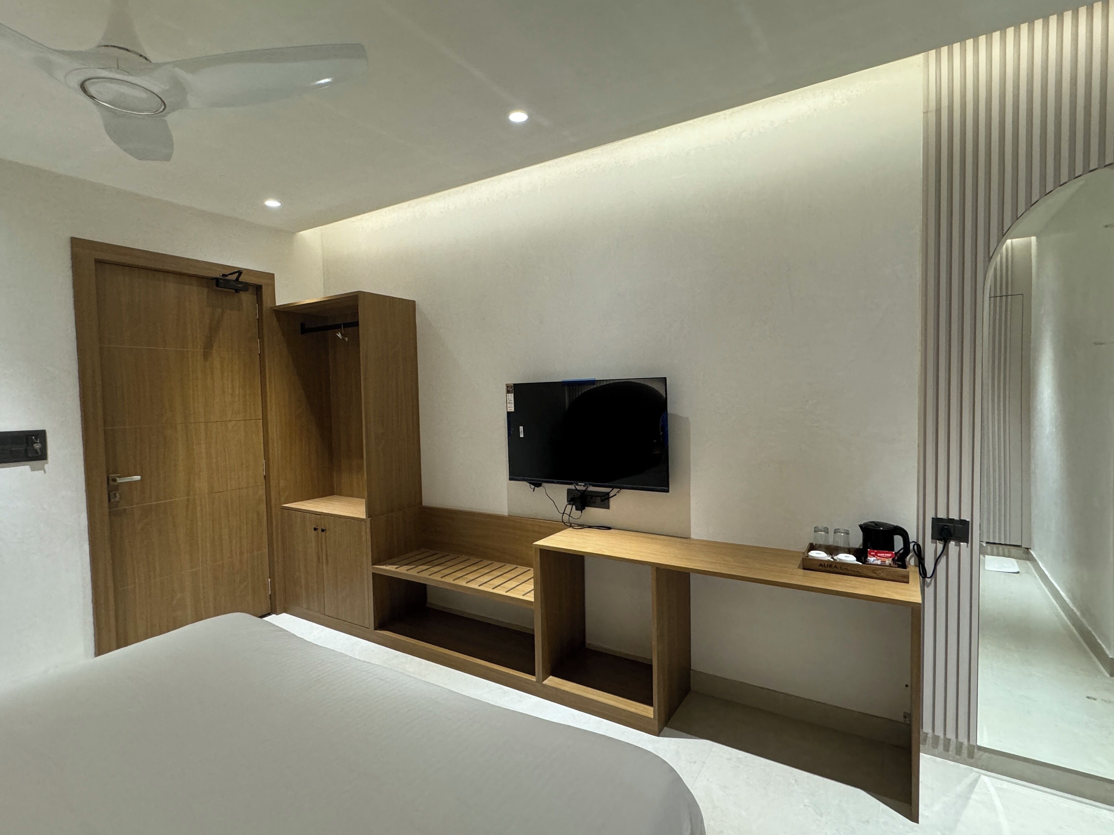 A modern hotel room with a bed, a TV mounted on the wall, a wooden wardrobe, and a desk with a mirror.