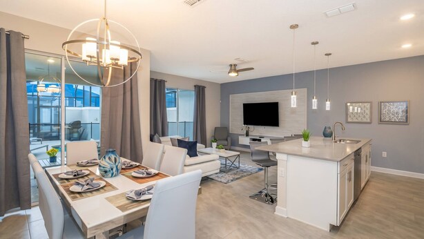 A modern kitchen with a central island, a dining area with a table set for four, and a living area with a sofa and TV. - Luxurious 4BR Home Pool Near Disney Storey Lake (Kissimmee)