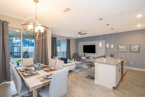 Una cocina moderna con una isla central, un comedor con una mesa preparada para cuatro personas y una sala de estar con sofá y televisor.