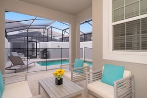 A covered outdoor seating area with a pool, white furniture, and a view of a house in the background.