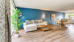 A living room with a blue brick wall, a white sofa, a wooden coffee table, and a dining area with chairs.