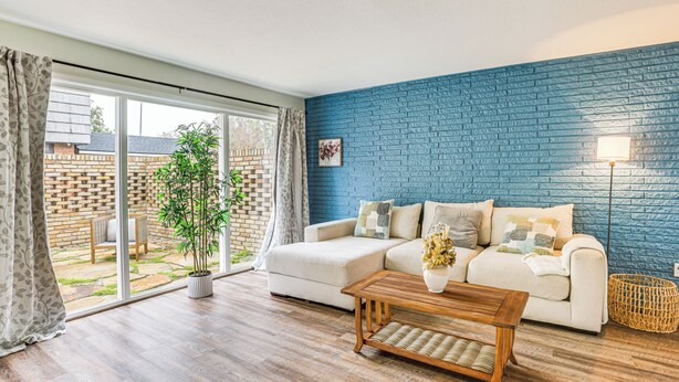 A living room with a blue brick wall, a white sofa, a wooden coffee table, and a large window with curtains.