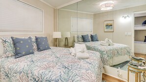 Quarto de hotel com duas camas, um espelho grande, uma mesinha e uma janela com persianas.