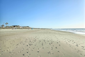 Une vaste plage de sable avec des empreintes, un bâtiment avec un toit visible au loin, et l’océan sur la droite.