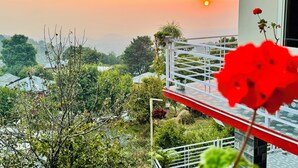 Een balkon met een rode bloem, met uitzicht over een tuin en huizen, met een zonsondergang op de achtergrond.