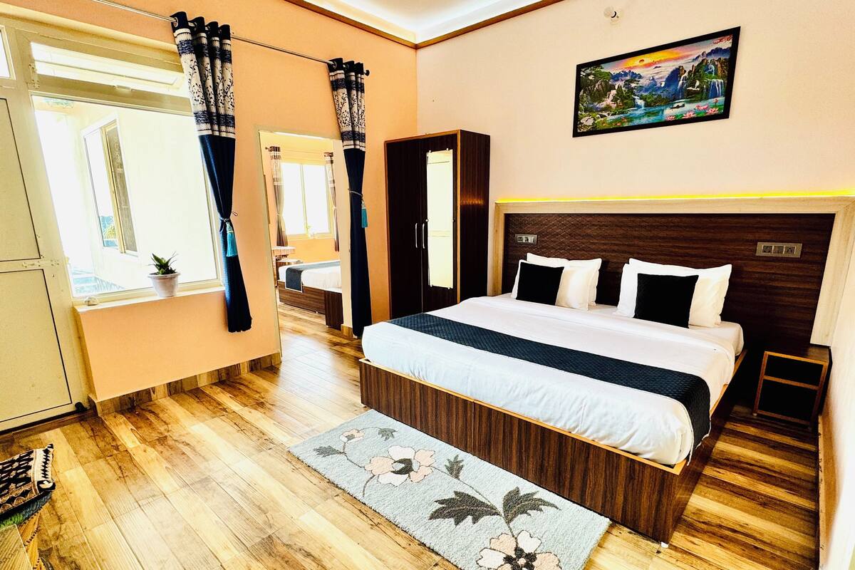 A hotel room with a large bed, wooden flooring, a small bedside table, a nightstand, a window with curtains, a door, and a painting on the wall.