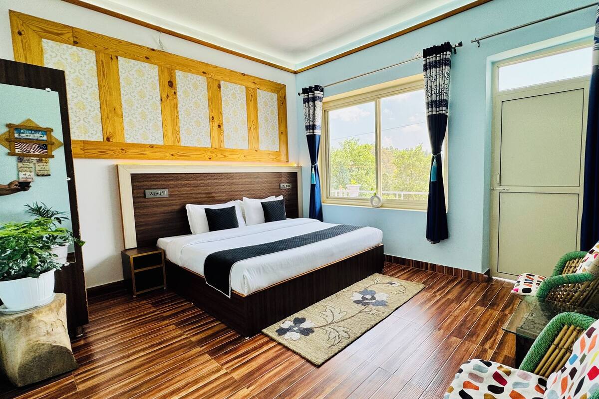 A bedroom with a wooden headboard, a large bed, a nightstand, a green chair, a window with curtains, and a view of trees.