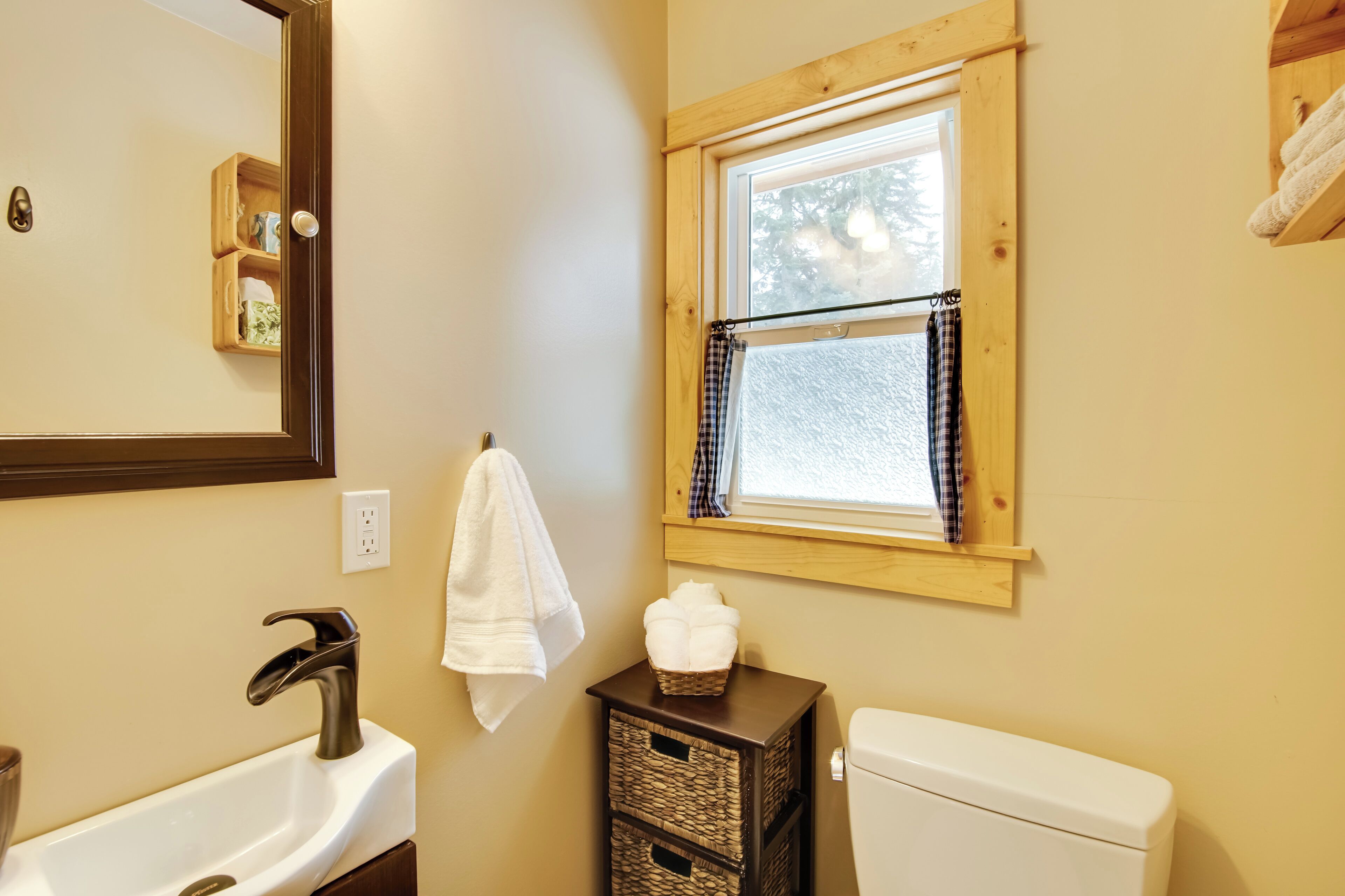 A bathroom with a white sink, a mirror, a white toilet, and a window with a plaid curtain.