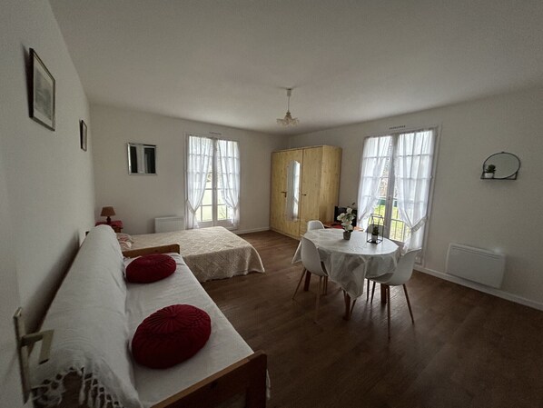 A spacious room with two single beds, a wooden wardrobe, a dining table with chairs, and large windows.