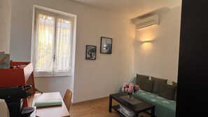 A living room with a sofa, a table with a vase, a window with curtains, and an air conditioning unit.