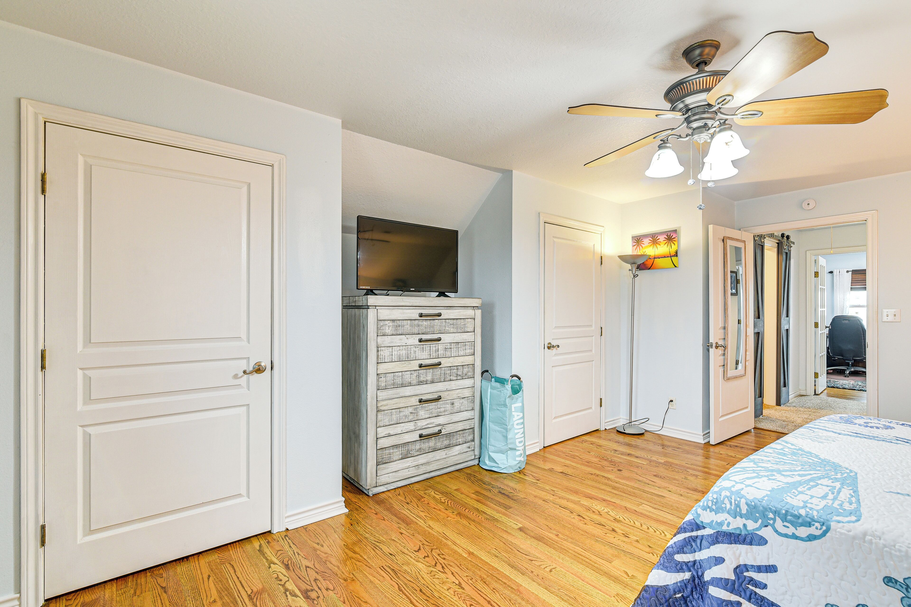 A bedroom with a wooden floor, a ceiling fan, a dresser, a television, and a door leading to another room.