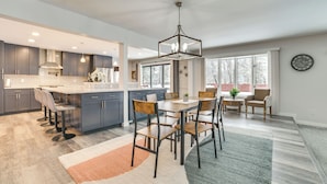 A modern kitchen with grey cabinets, a central island, and bar stools. There is a dining area with a table and chairs, and a large window providing natural light. - 3 Mi to Dtwn Alpena: Family Home w/ Yard! (Alpena)