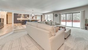 A spacious living room with a large beige sectional sofa, a kitchen area with dark cabinets and a central island, and large sliding glass doors leading to a snowy outdoor area. - 3 Mi to Dtwn Alpena: Family Home w/ Yard! (Alpena)