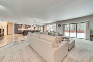 Amplia sala de estar con un sofá seccional beige grande, una zona de cocina con armarios oscuros y una isla central, y grandes puertas correderas de vidrio que dan a un área exterior nevada.