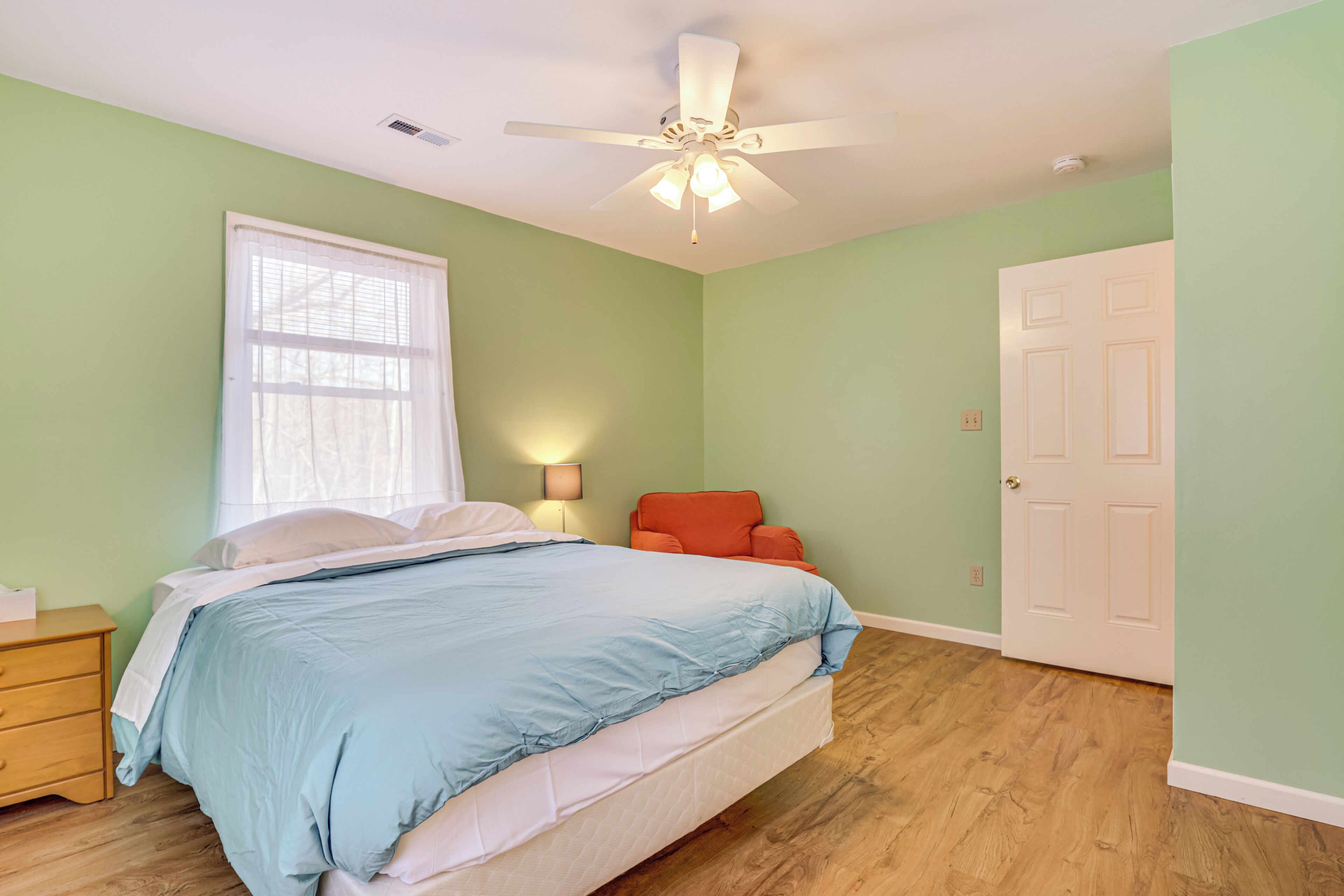 Une chambre à coucher comprenant un lit, un ventilateur de plafond, une fenêtre avec des rideaux, une table de chevet, une chaise et une porte.