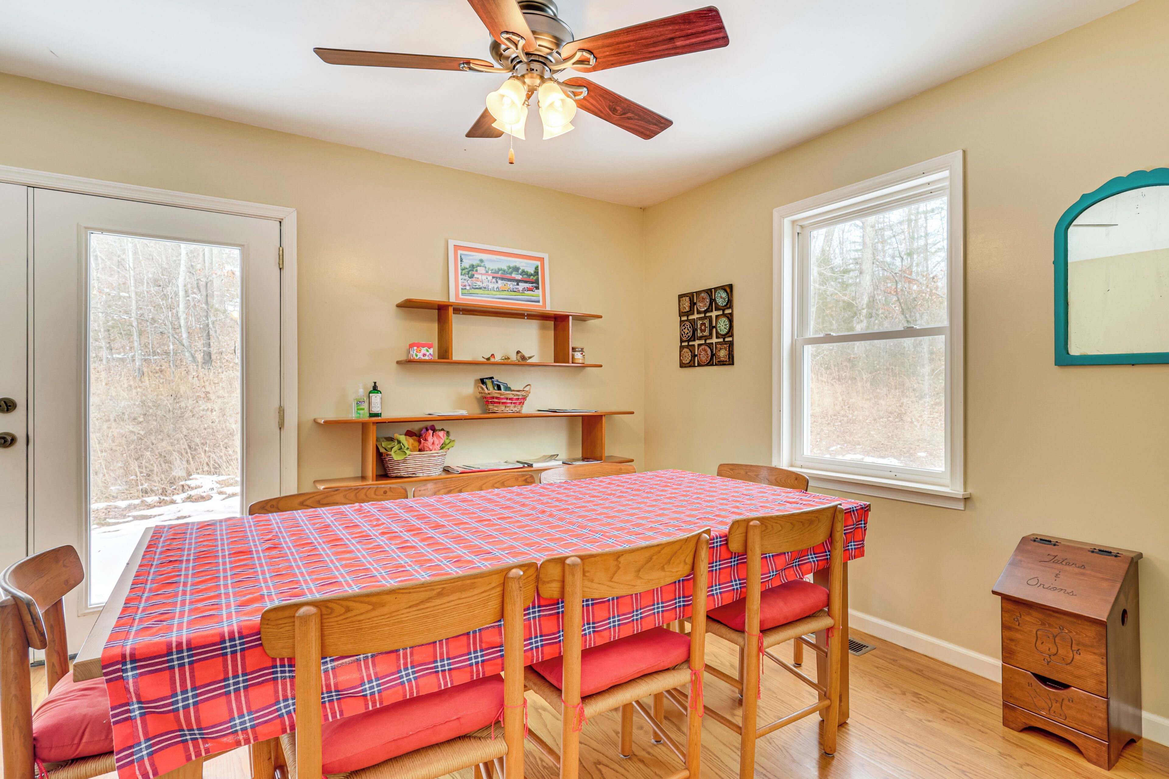 A dining area with a plaid tablecloth, wooden chairs, a ceiling fan, and a window with a view of trees.