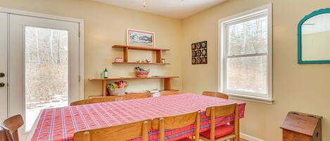 A dining area with a plaid tablecloth, wooden chairs, a ceiling fan, and a window with a view of trees.