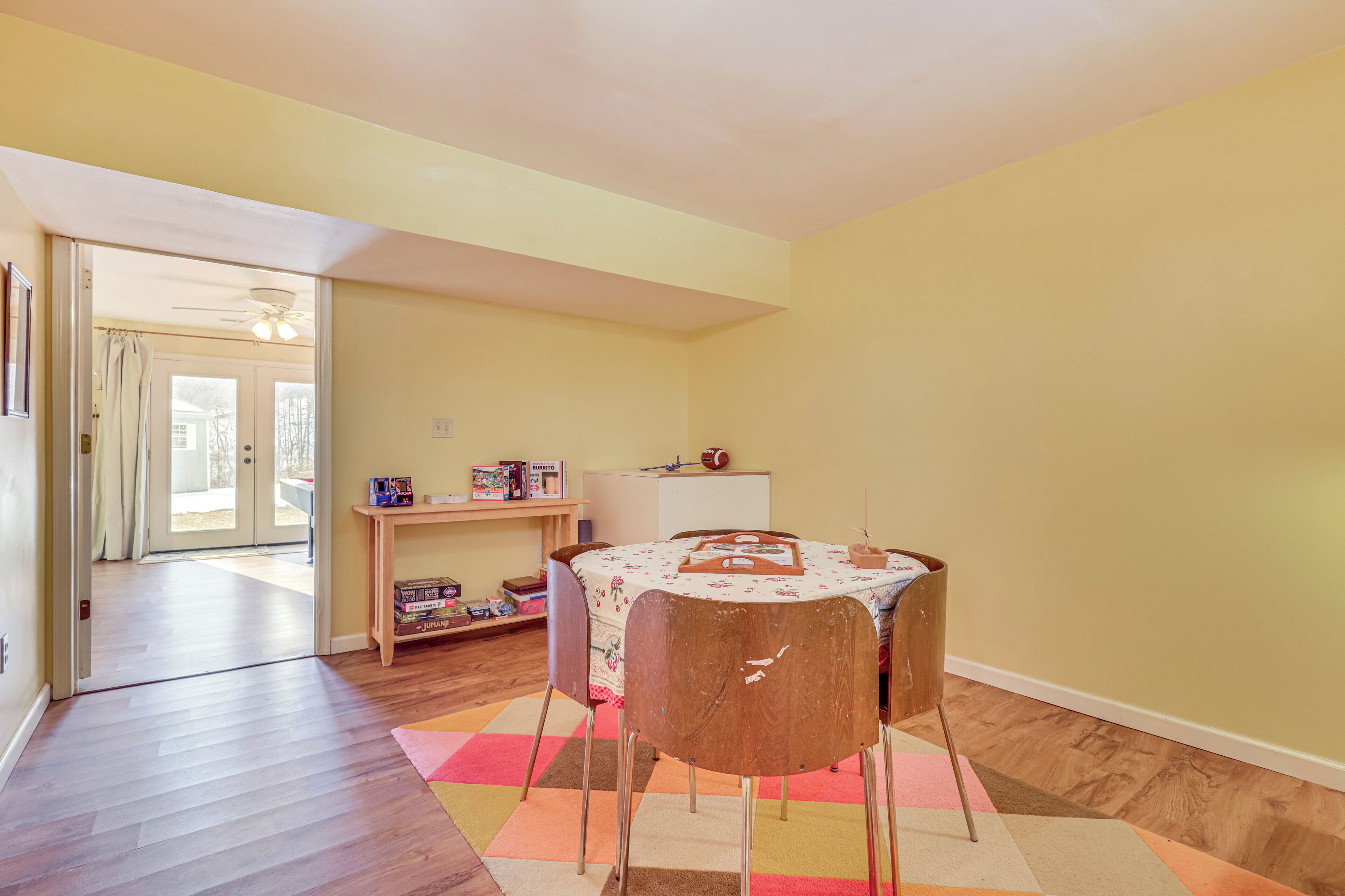 A dining area with a round table, chairs, and a colorful rug. There is a door leading to another room and a small shelf with toys.