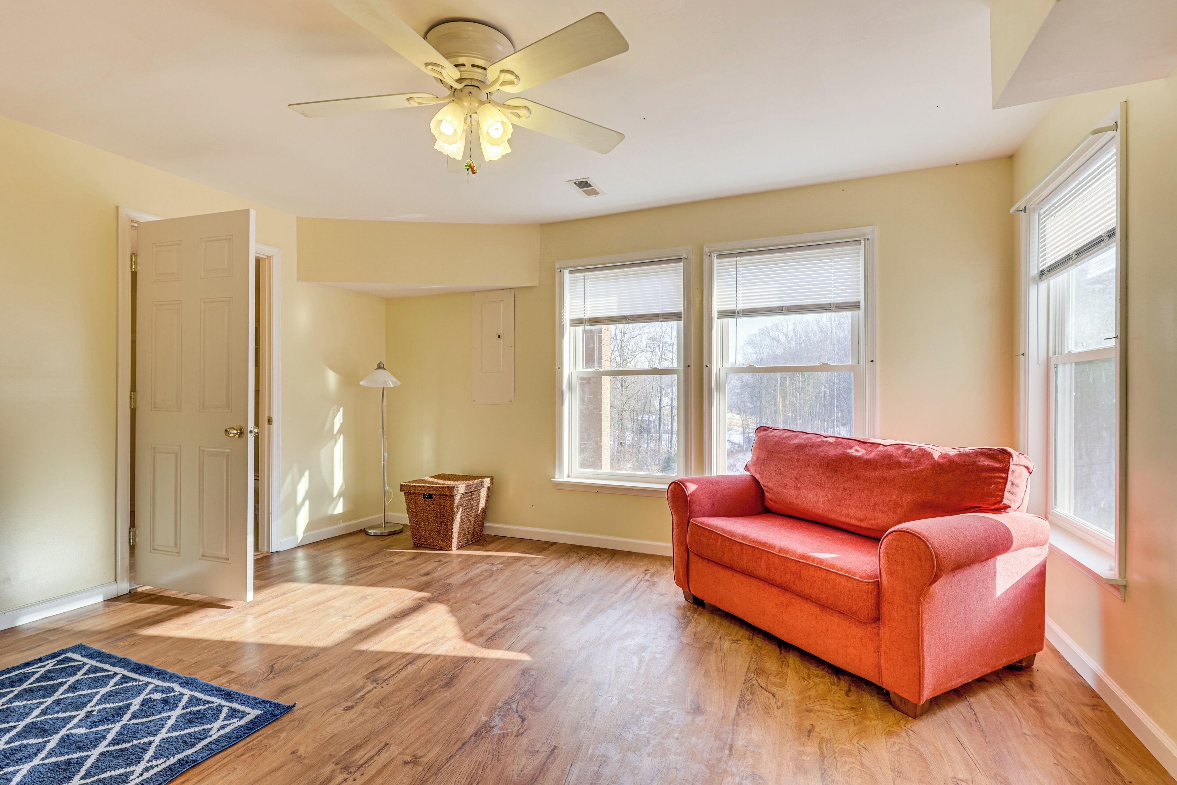 A room with hardwood flooring, a red sofa, a ceiling fan, and two windows with blinds.