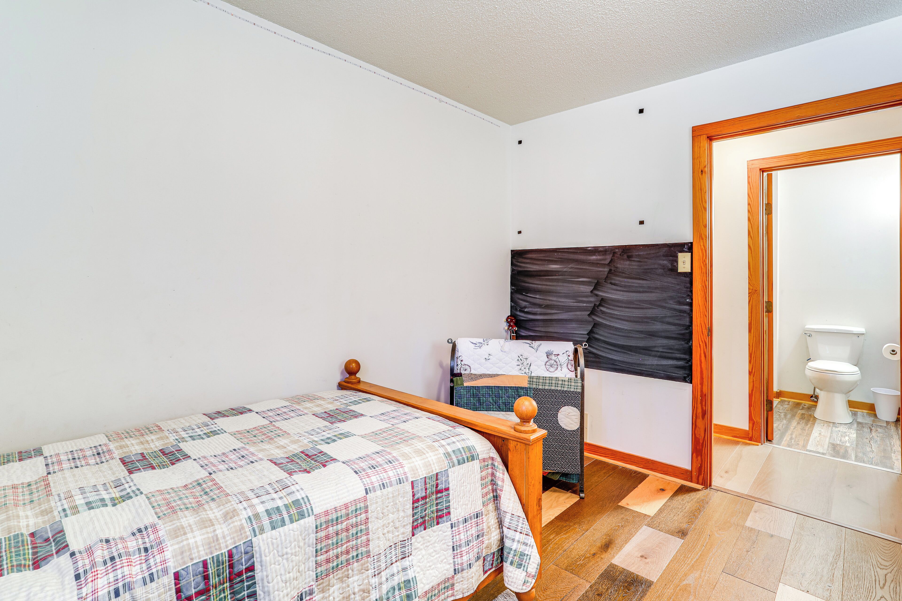 Un dormitorio pequeño y ordenado con una cama individual, una silla y un baño visible a través de una puerta abierta.