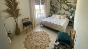 Ein Schlafzimmer mit Bett, Deckenventilator, großem Fenster mit Vorhängen, Wandgemälde, Teppich und einem Sessel.