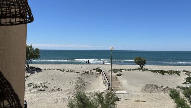Ein Strandblick mit einem sandigen Pfad, der zum Wasser führt, einem Laternenpfosten und vereinzelter Vegetation.